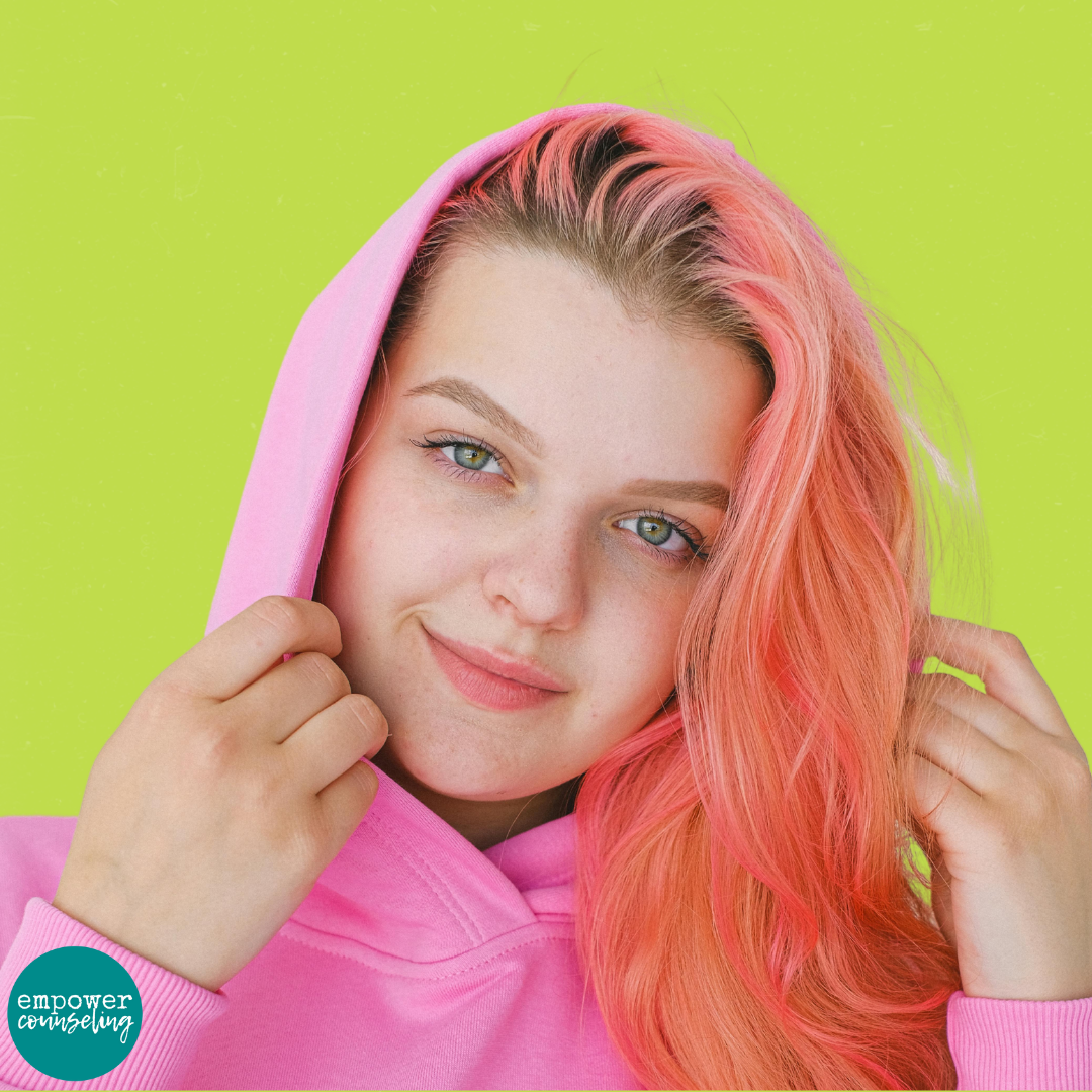 Teen girl with pink hair wearing a pink hoodie looks into the camera with a confident expression, standing against a bold chartreuse background. What parents need to know about teen therapy.